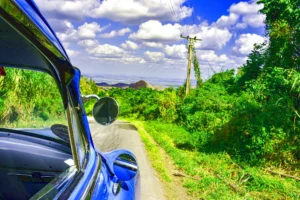 "Vintage blue car driving through a scenic green countryside road with distant hills and a bright blue sky with clouds"
