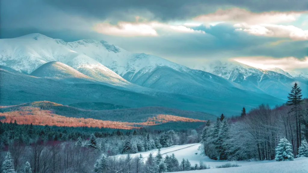 Snow-covered mountains and forests beneath a partly cloudy sky, with sunlight illuminating parts of the landscape and shadows cast on the trees and hills.